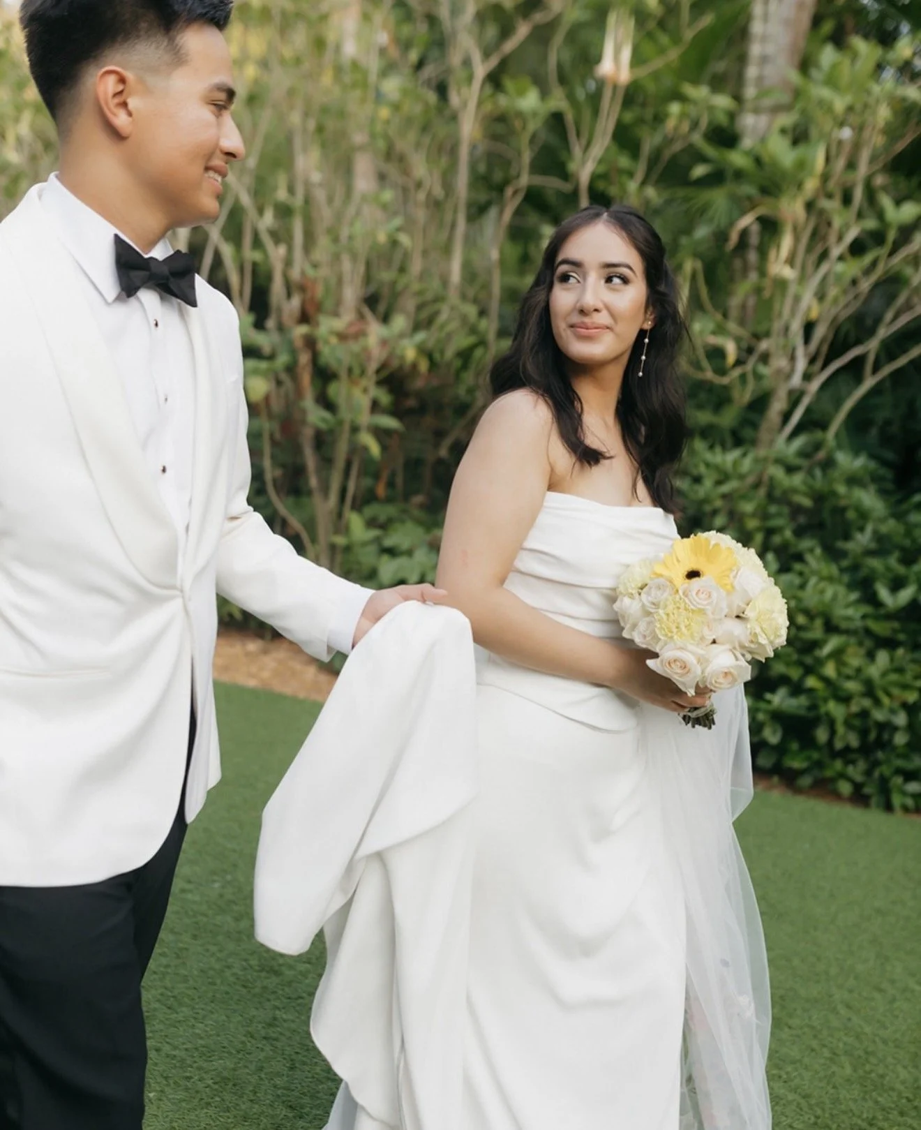 Groom in white tuxedo jacket helping bride in strapless white gown walk while she looks back and smiles, holding a yellow and white bouquet, at Sunken Gardens Tampa wedding planned by Peony Wedding Co.