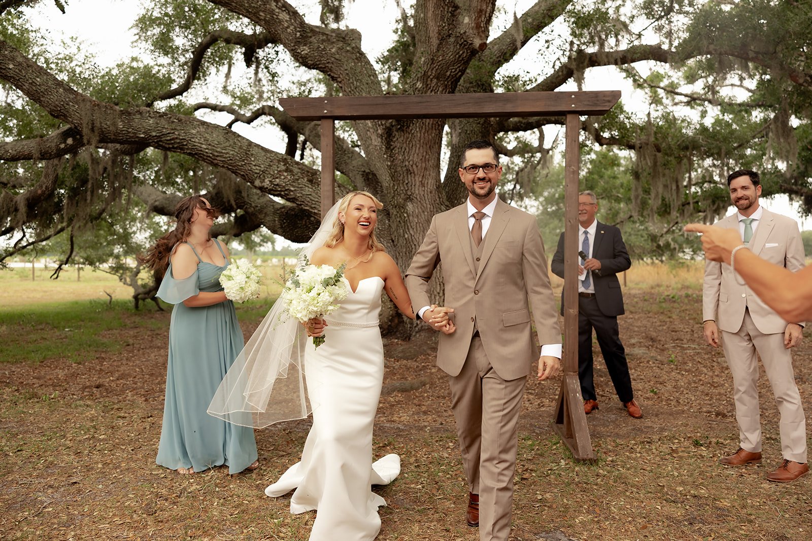 Joyful bride and groom walking back down the aisle after the ceremony at Browns Grove Sarasota, surrounded by wedding party under oak tree