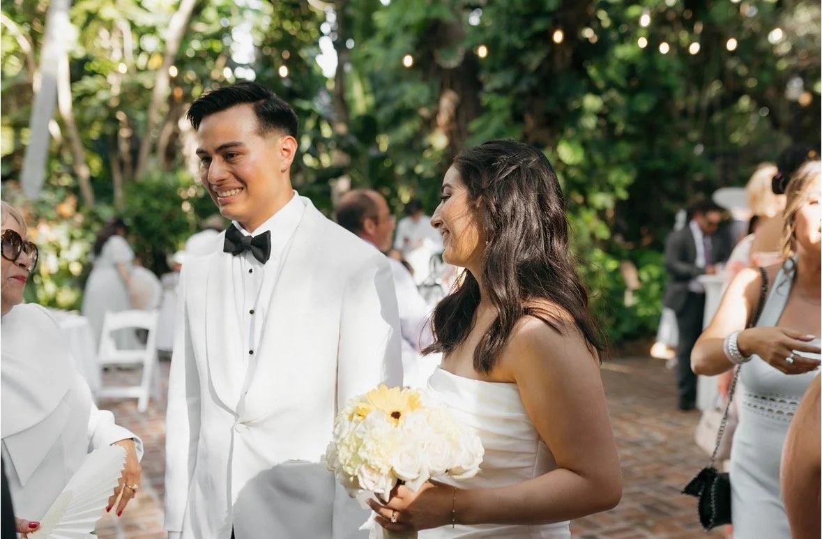 Bride and groom walking the recessional through a tropical tree canopy with string lights overhead at Sunken Gardens Tampa wedding, surrounded by smiling guests, planned by Peony Wedding Co.