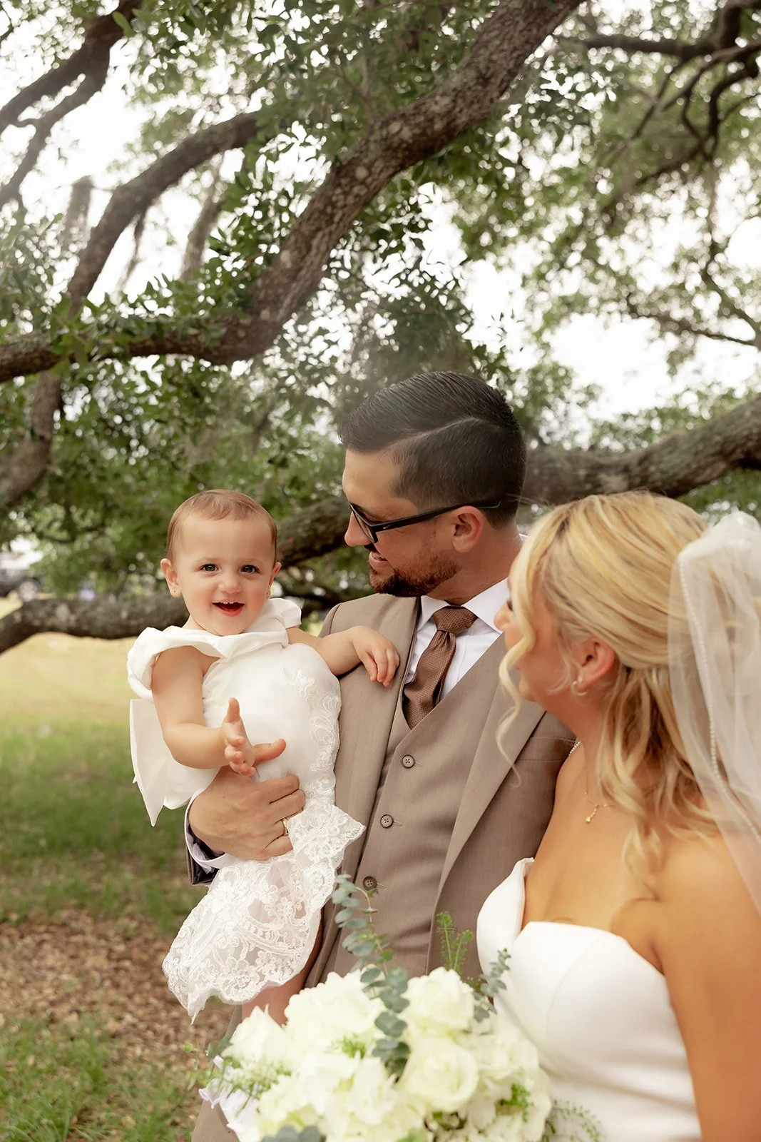 Bride, groom, and baby daughter in lace dress under the live oak tree at Browns Grove Sarasota — family portrait