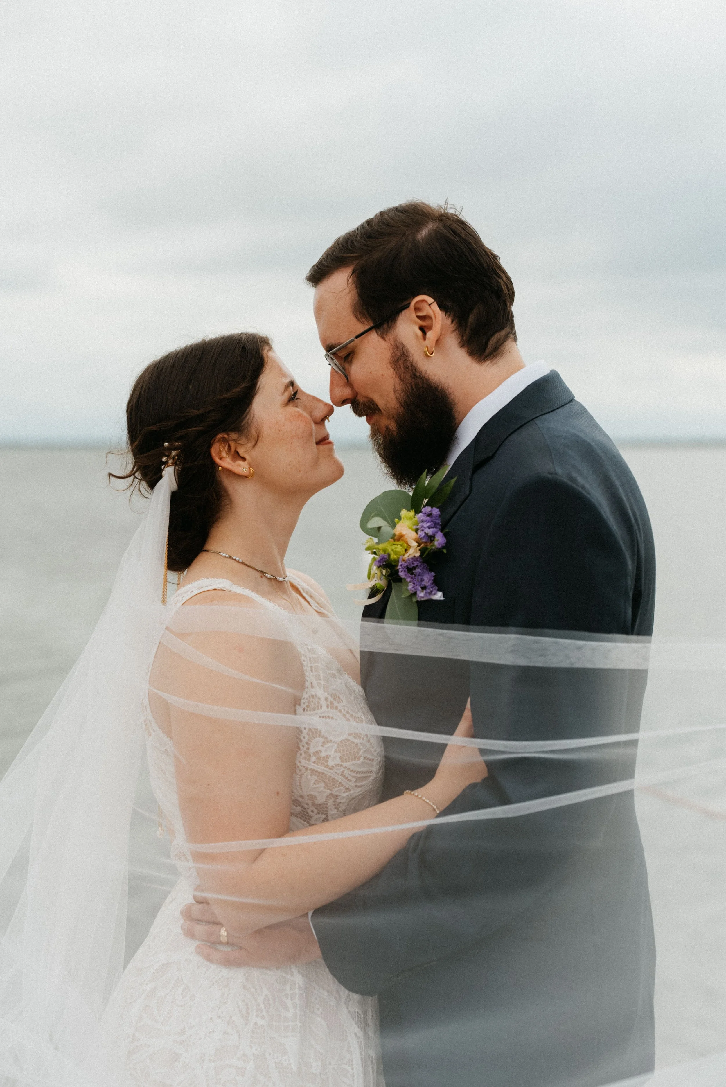 Bride in a lace wedding gown and long veil touching foreheads with her groom in an intimate outdoor moment, with a soft grey sky behind them.