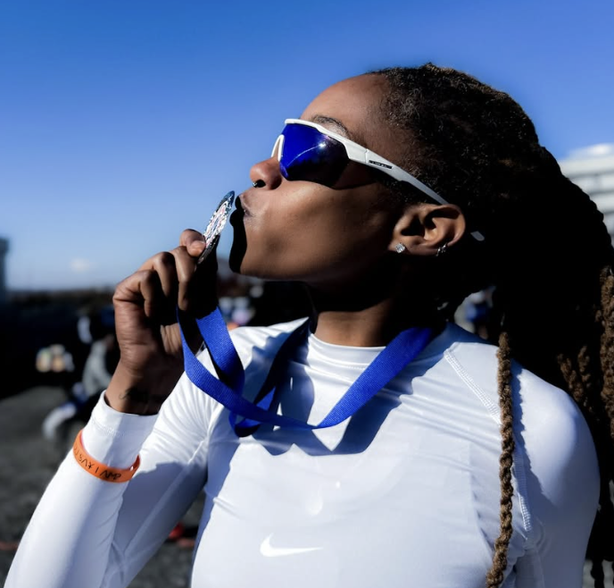 Female athlete with long dreadlocks wearing sunglasses and a white sports shirt, holding a silver medal and kissing it, with a clear blue sky background.