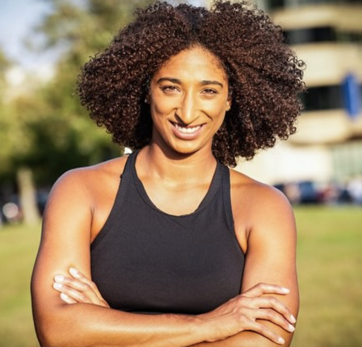 A woman with brown, curly hair smiling outdoors with arms crossed, wearing a black athletic tank top, on a sunny day.