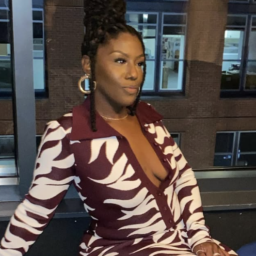A woman with braided hair wearing a purple and white patterned dress with a deep neckline, standing outside a building at night.