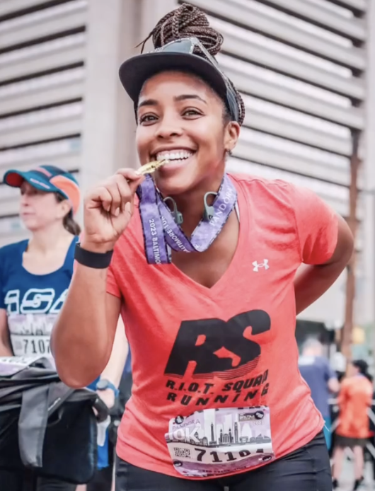 Woman with braided hair wearing an orange running shirt, holding a medal, smiling after a race.