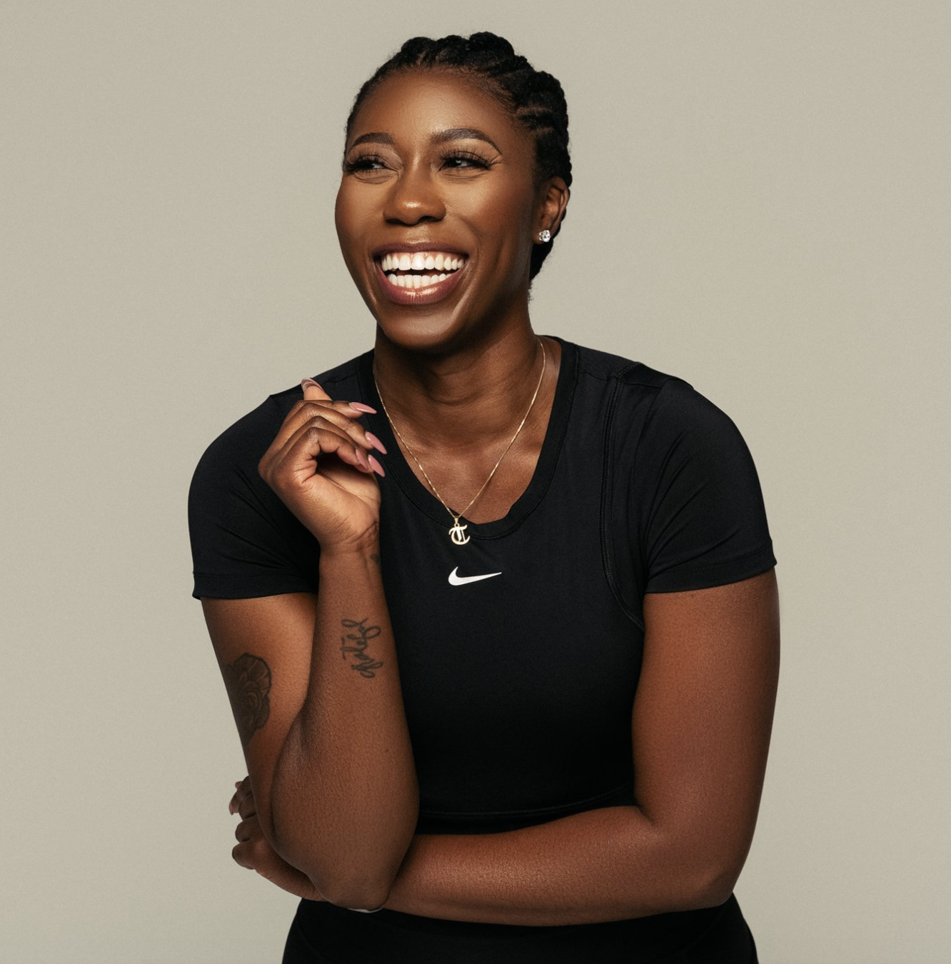 Smiling woman with braided hair wearing a black Nike athletic shirt, gold necklace, and earrings, against a plain background.