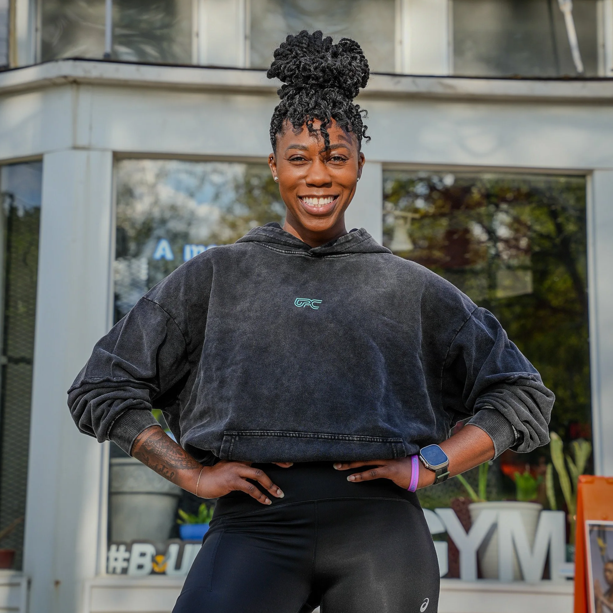 A woman with short dark hair, smiling, wearing a white tank top, standing against a gray textured wall.