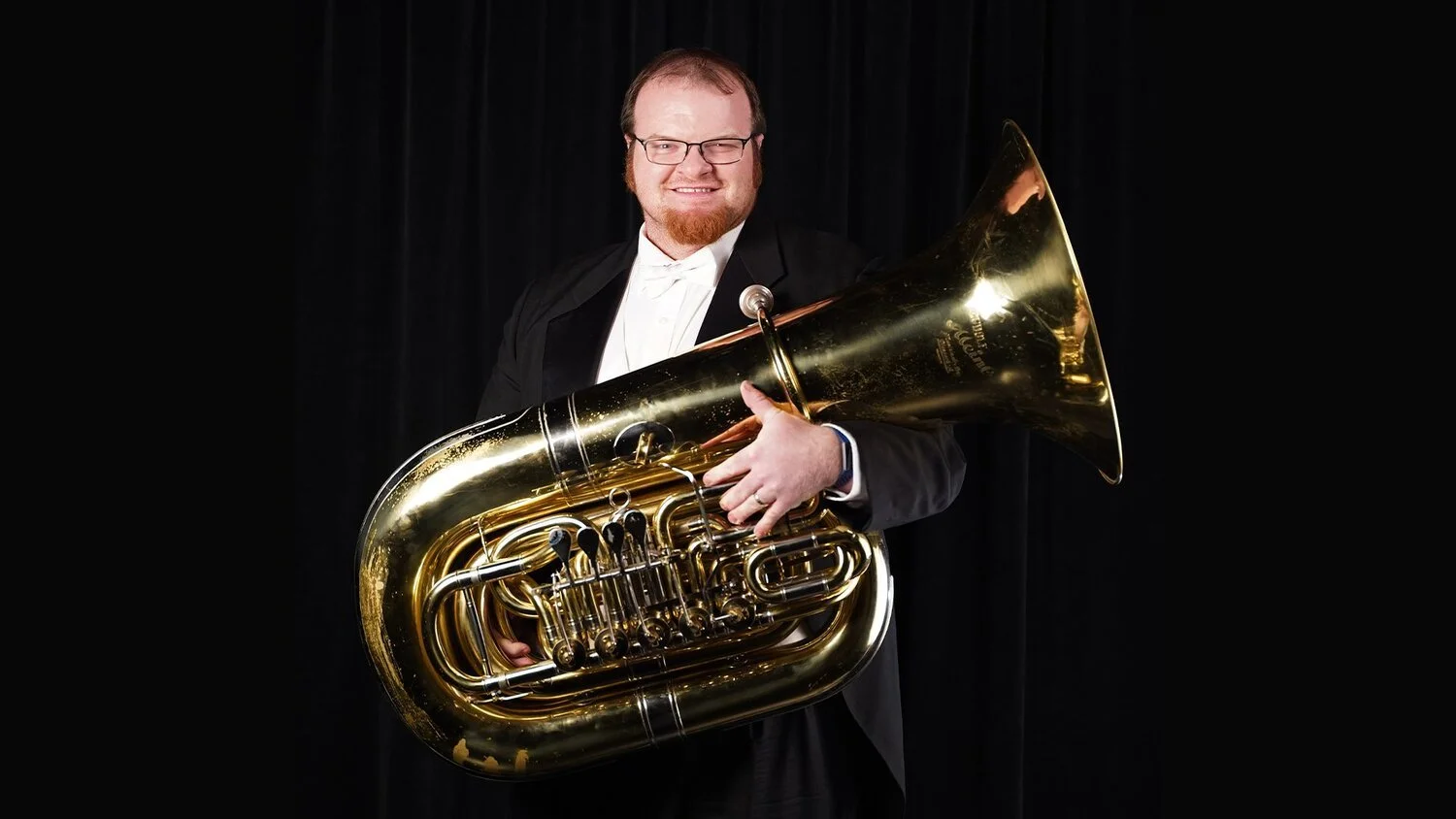A man with glasses and a beard, wearing a tuxedo, holds a large brass tuba and smiles at the camera against a black background.