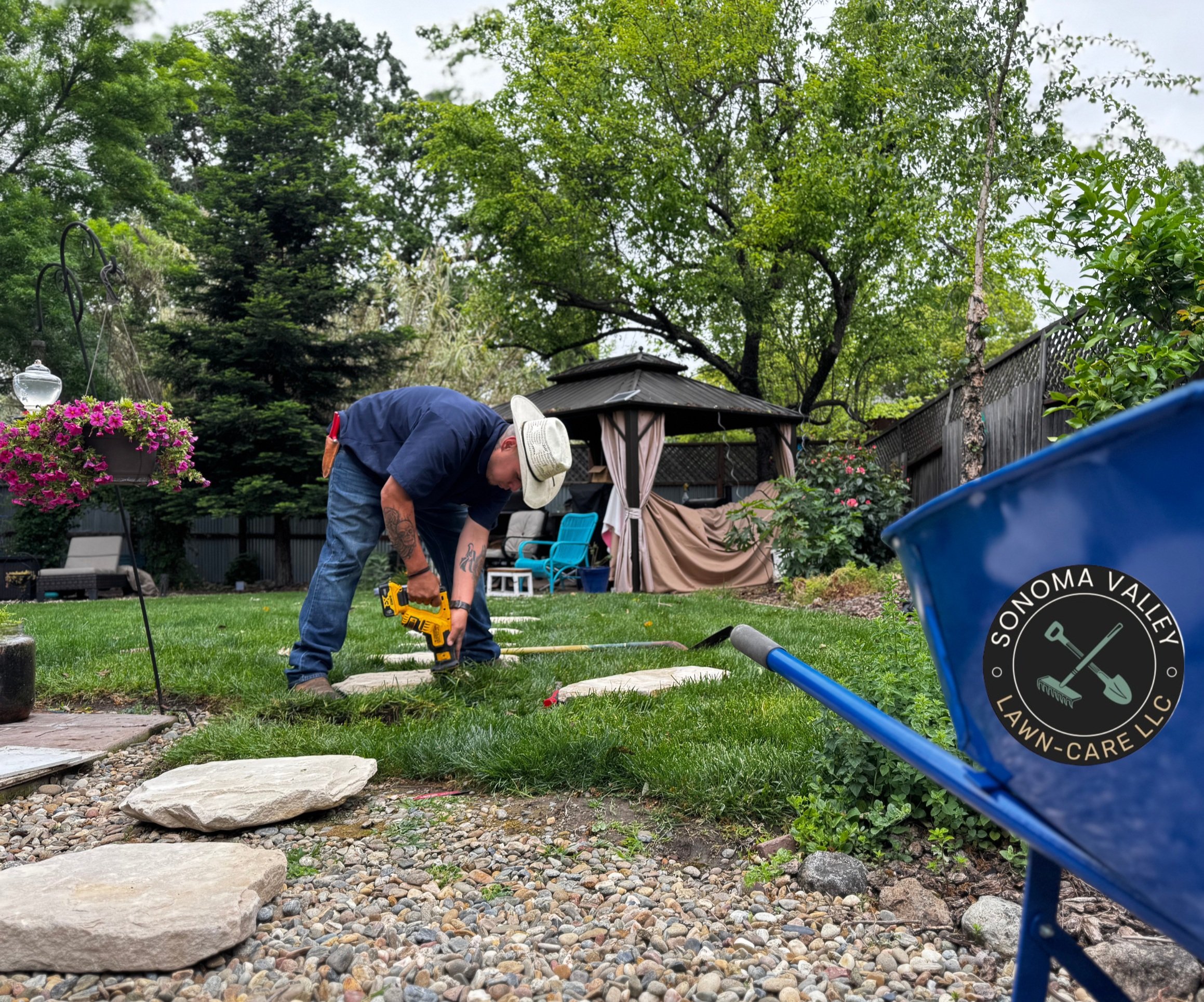 A person wearing a hat is using a power tool on a stepping stone in a backyard with a gazebo. The garden has trees, flowers, and garden furniture. A blue wheelbarrow with a "Sonoma Valley Lawn-Care LLC" logo is in the foreground.