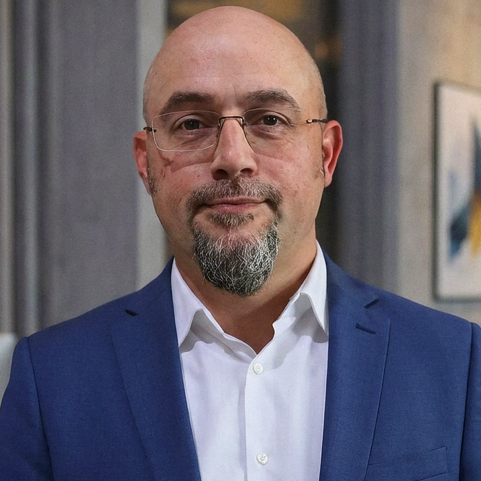 Close-up of a bald man with glasses, a goatee, wearing a blue suit jacket and a white dress shirt, standing indoors.
