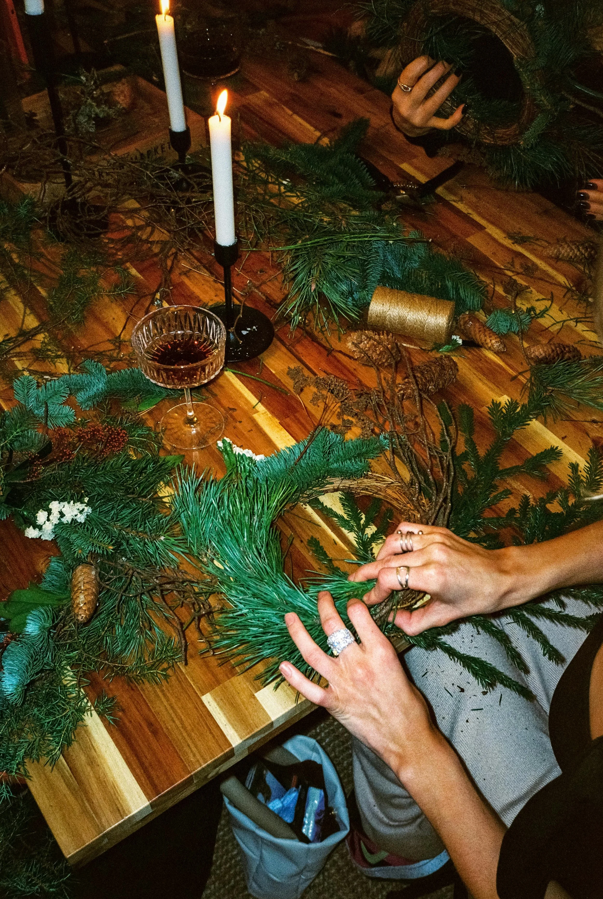 People decorating a holiday wreath with pine branches, pinecones, and other natural elements on a wooden table, with lit candles and a glass of red wine nearby.