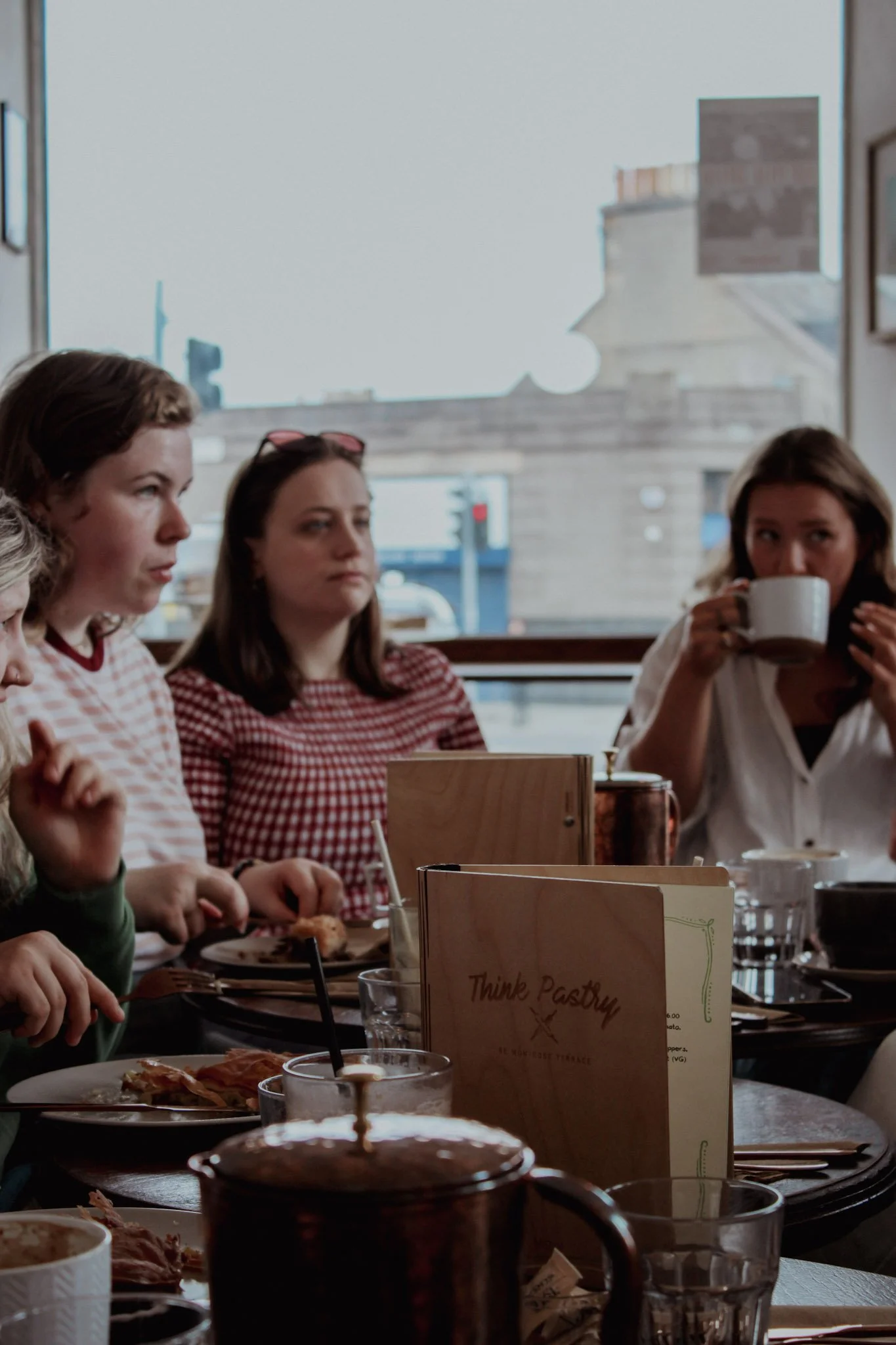 Four women seated at a table in a cozy cafe with large windows, eating and drinking, with a menu titled "Think Pastry" on the table.