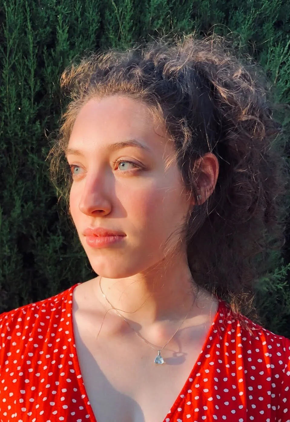 A young woman with curly hair and fair skin wearing a red polka dot dress and a necklace, standing outdoors against green foliage, bathed in warm sunlight.