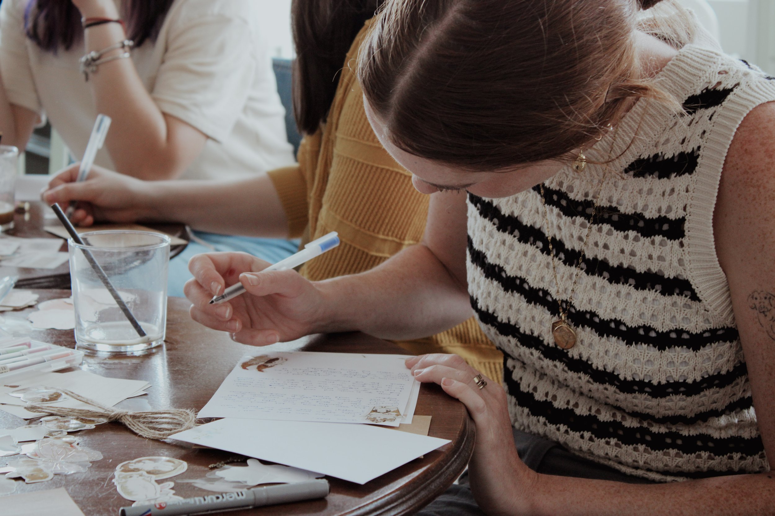 A person with shoulder-length brown hair wearing a black and white striped sleeveless top is writing on a piece of paper with a white marker at a table. There are various crafting supplies, including a glass of water with a paintbrush, scattered on the table.