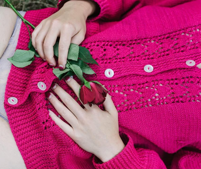 A person in a pink knitted sweater holds a single red rose with green leaves.
