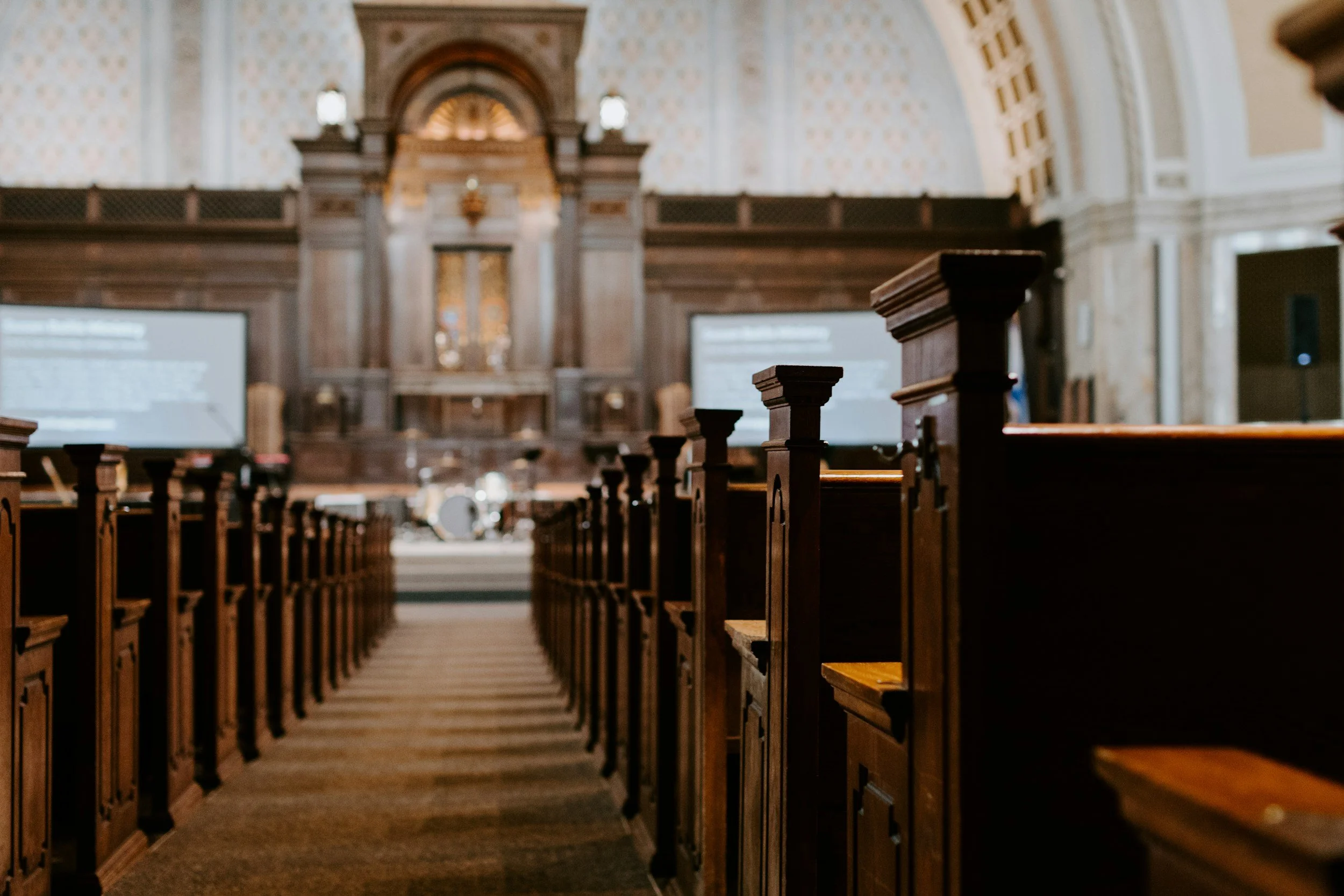 Inside a church with wooden pews on either side leading to an altar with ornate woodwork and decorative patterns on the wall behind.