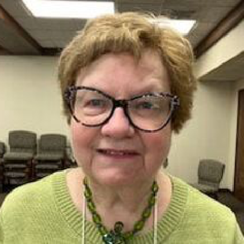 A woman with short, light brown hair wearing glasses and a green necklace, sitting in a room with stacked chairs and a wooden ceiling