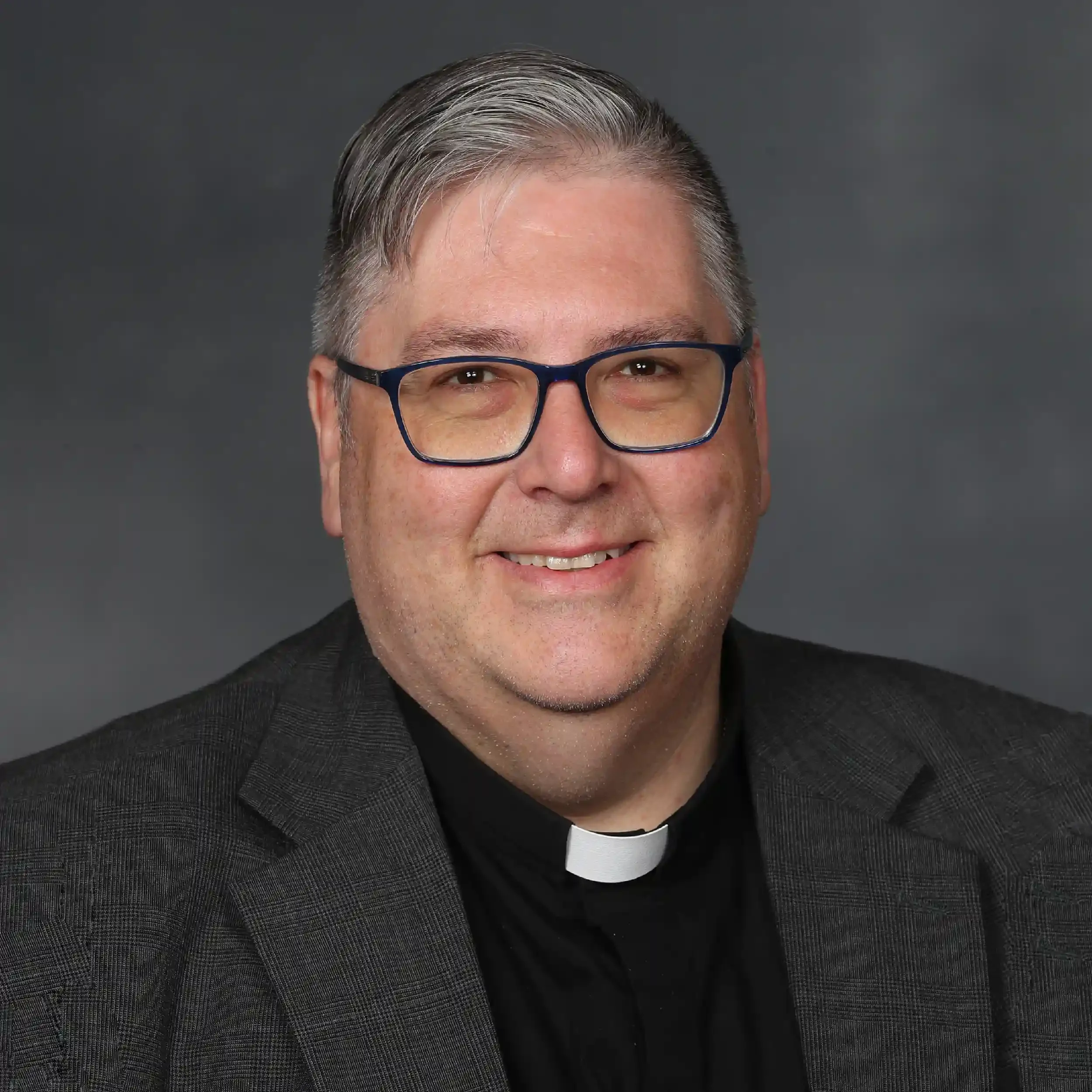 Close-up of a smiling man wearing glasses and a black clerical collar, dressed in a dark suit, against a dark gray background.