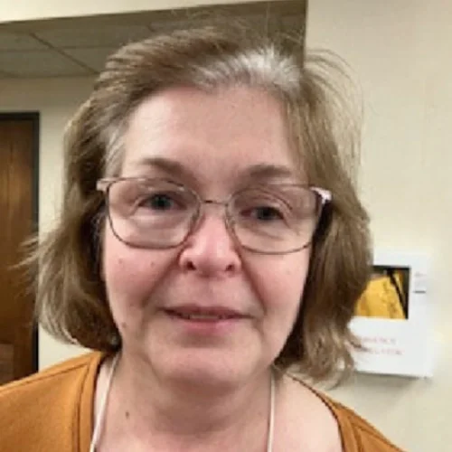 A middle-aged woman with short, light brown hair wearing glasses and an orange top indoors.