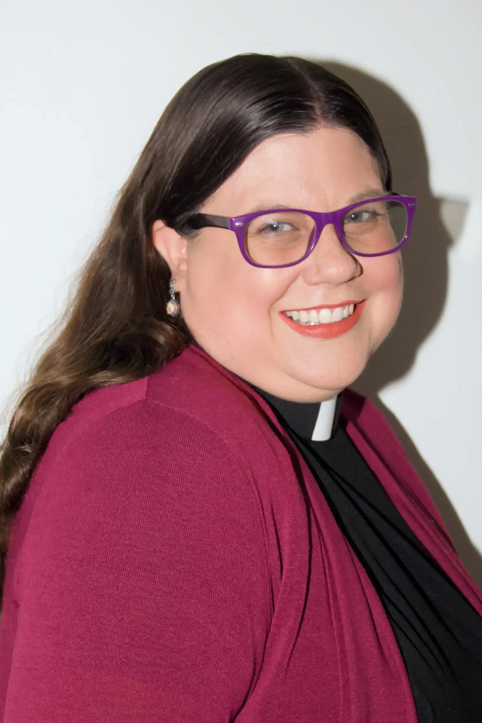 Close-up of a woman with long brown hair, wearing purple glasses, a dark top, and a maroon blazer, smiling in front of a plain white background.