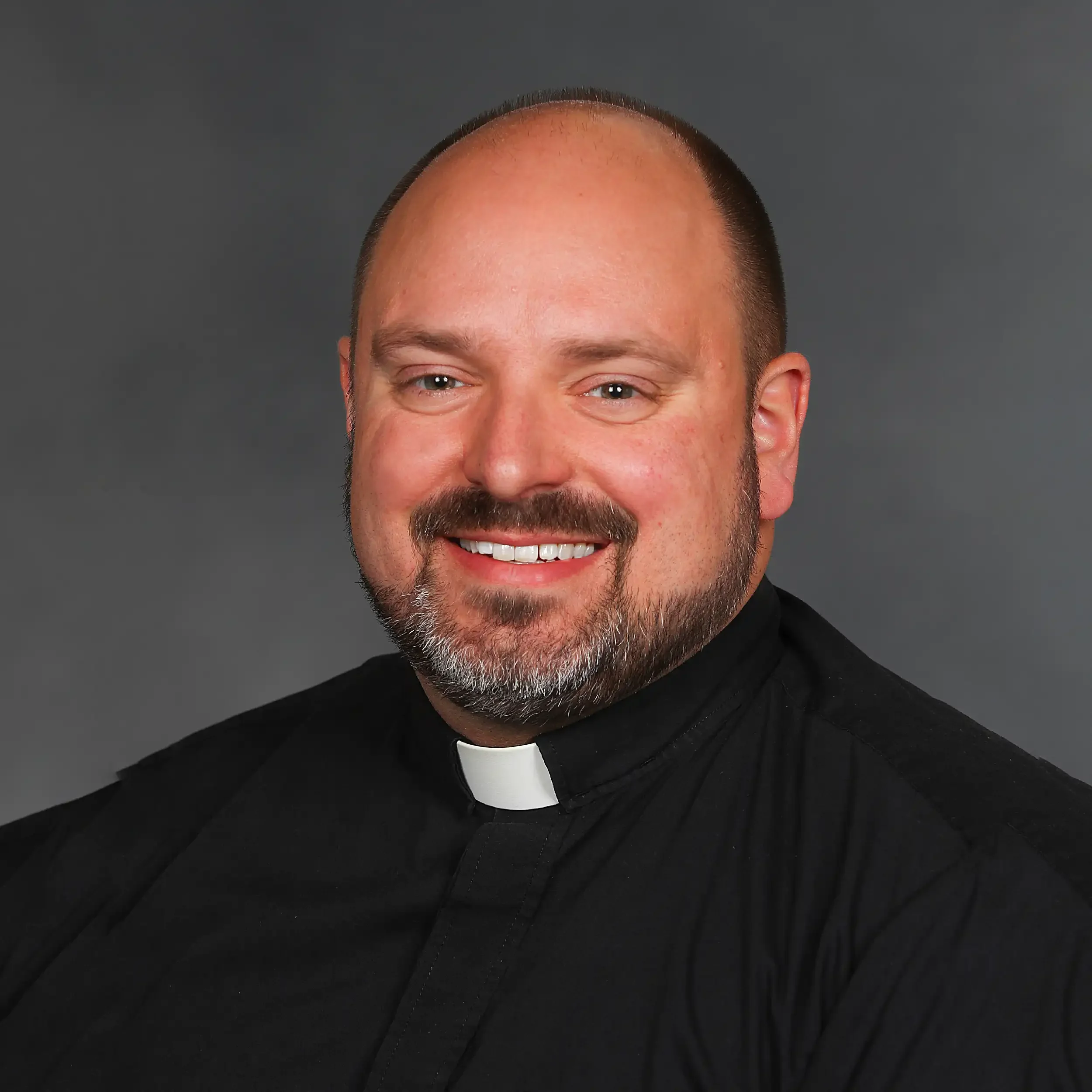 A man with a light beard and mustache wearing a black clerical shirt with a white clerical collar, smiling against a plain gray background.