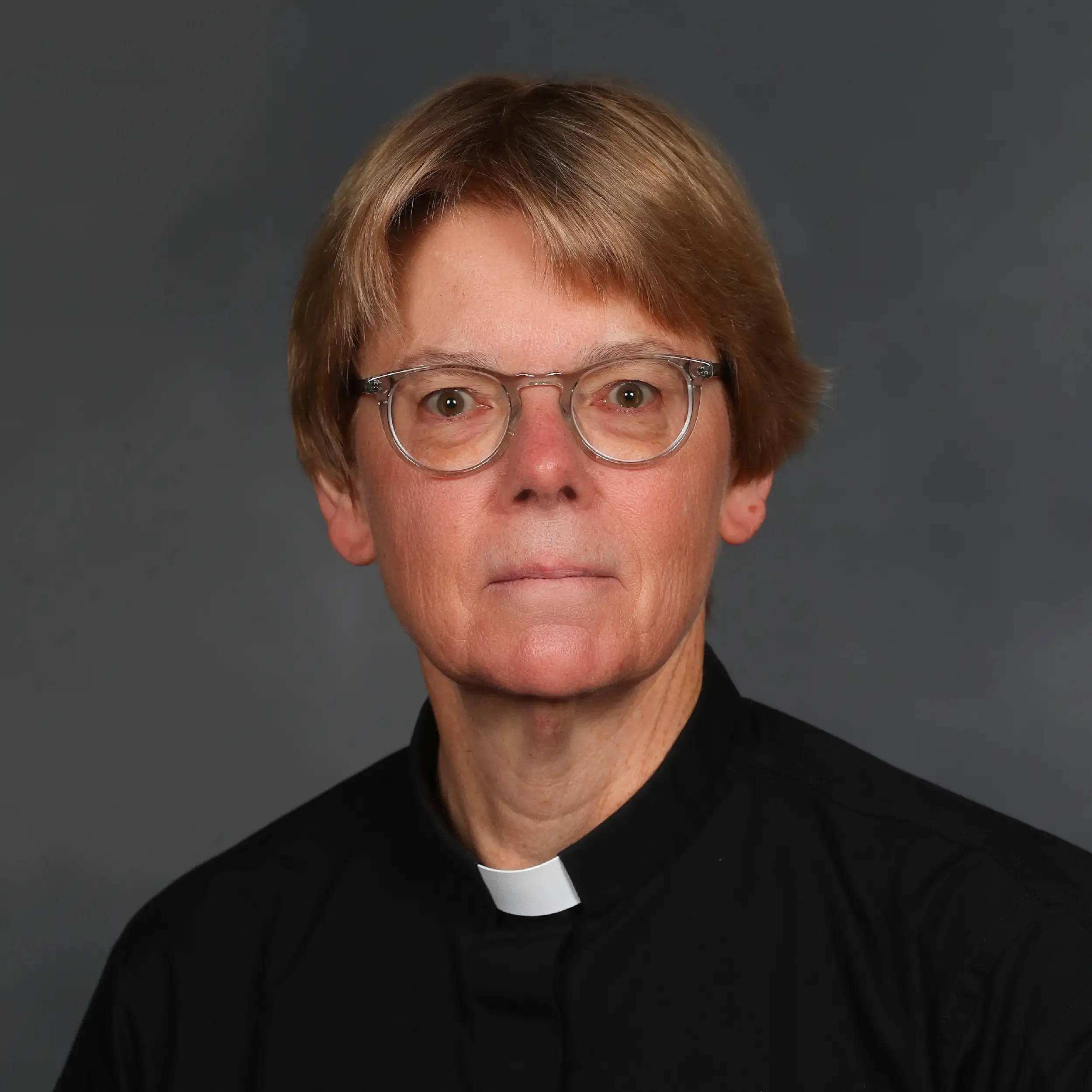 A person with light brown hair wearing glasses and a black clergy shirt with a white clerical collar.