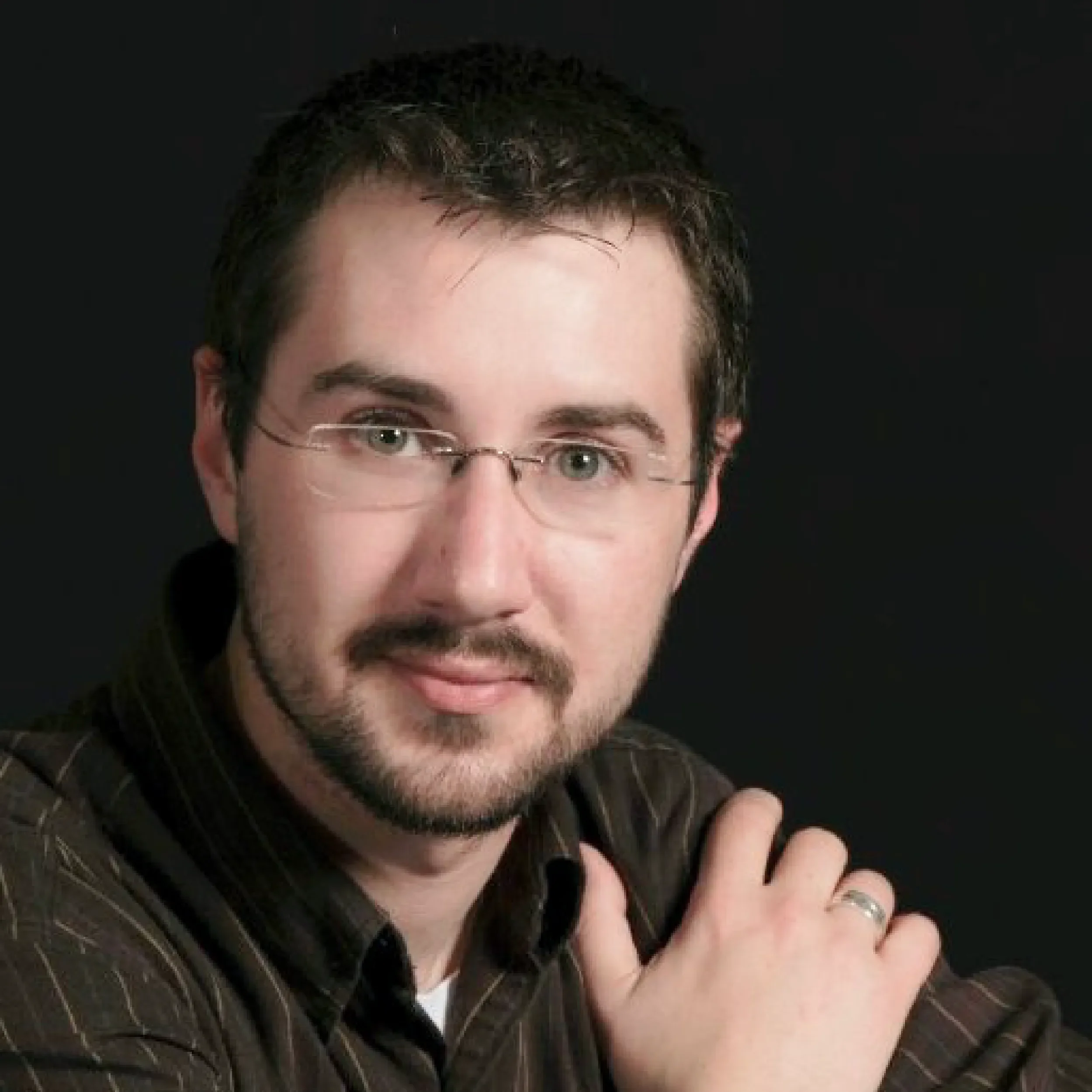 Close-up of a person's face, showing part of their forehead, eyes, nose, and lips. They have short dark hair, glasses, and facial hair, with a dark background.