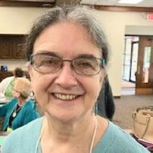A smiling woman with glasses and gray hair indoors, with other people and wooden furniture in the background.