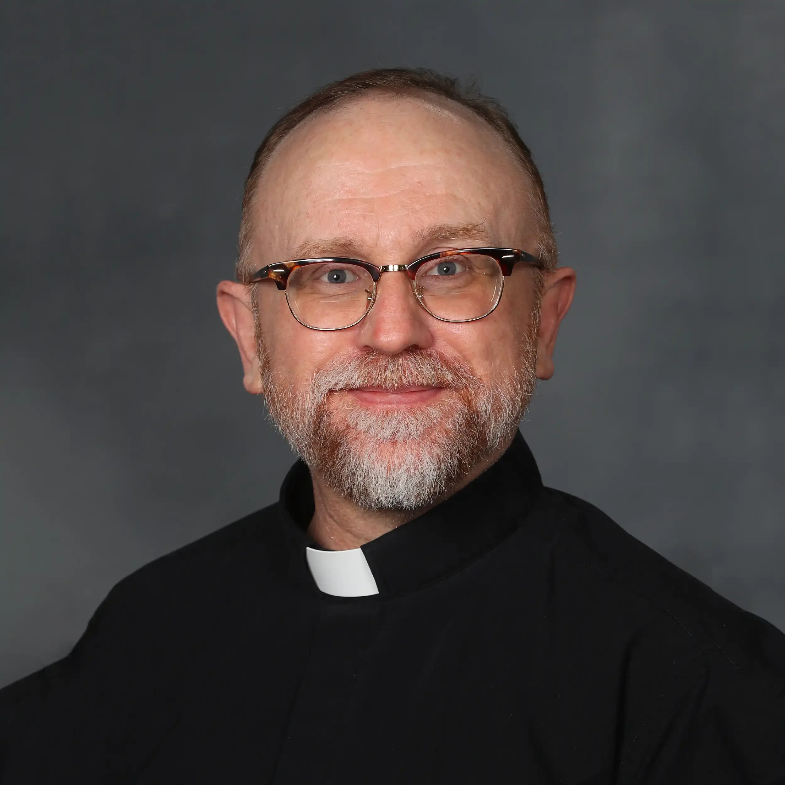 Close-up of a man wearing glasses and a clerical collar, smiling, against a plain gray background.