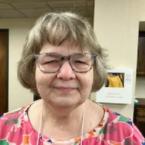 A smiling elderly woman with glasses and short gray hair, wearing a colorful patterned shirt, standing indoors.