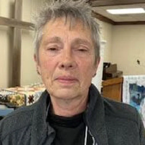 An older woman with short, gray hair and a serious expression, standing indoors near a wooden structure and decorated with patterned fabric.