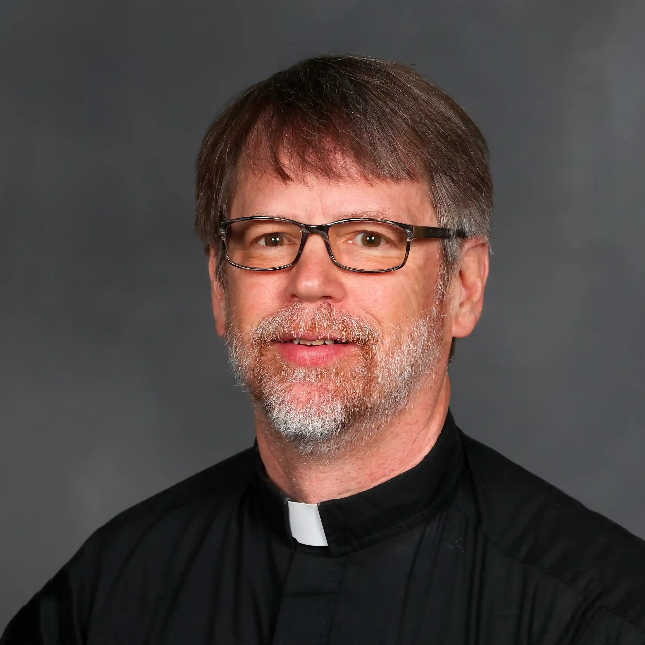 Close-up of a middle-aged man with light skin, gray hair, and glasses dressed as a priest, wearing a clerical collar, against a dark gray background.