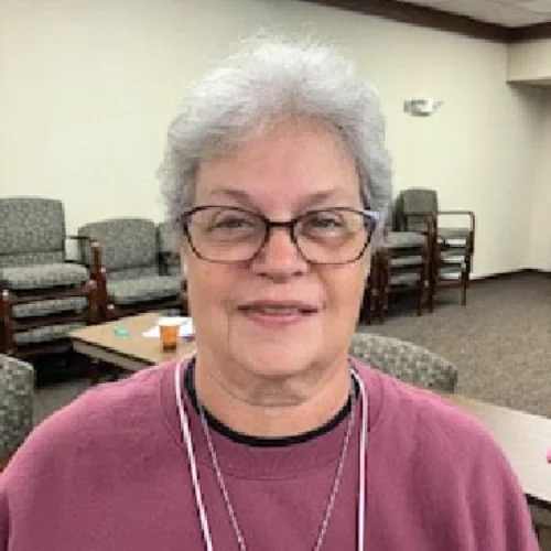 An older woman with short gray hair, glasses, and a pink sweatshirt, smiling in a room with chairs.