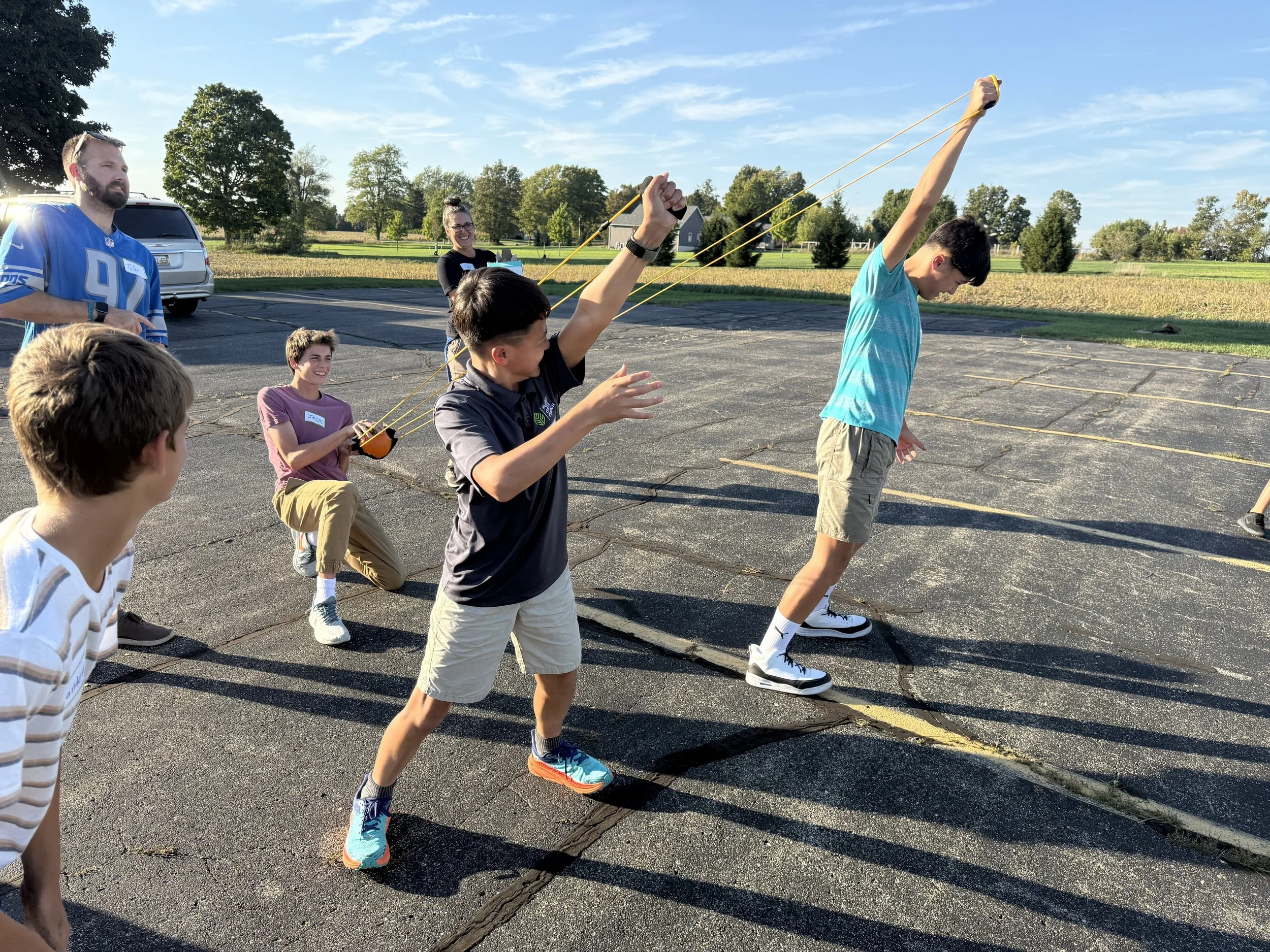 Children and adults participating in an outdoor activity involving stretching and using resistance bands, on a parking lot surrounded by green fields and trees under a blue sky.