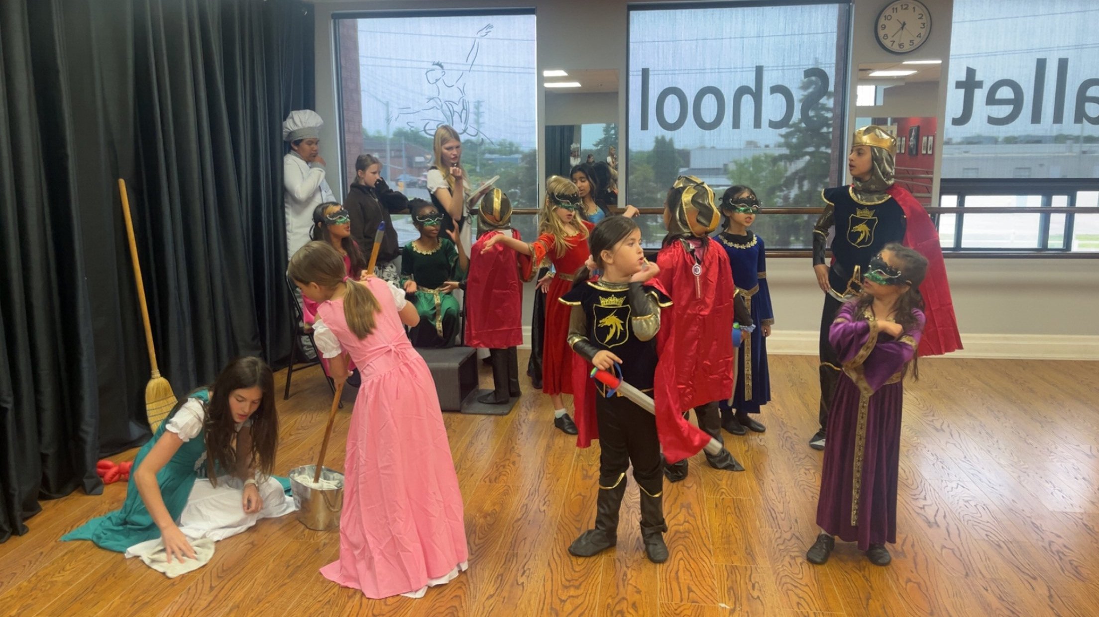 Children dressed as medieval knights and princesses practicing a play or costume activity in a room with a wooden floor, along with a person dressed as a chef and a woman in white. Some children are wearing masks, capes, and armor, with a window in t