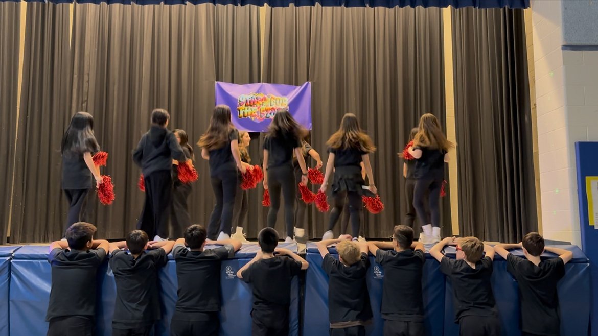Group of performers on a stage holding red pom-poms, with a purple banner in the background, and children watching from below.