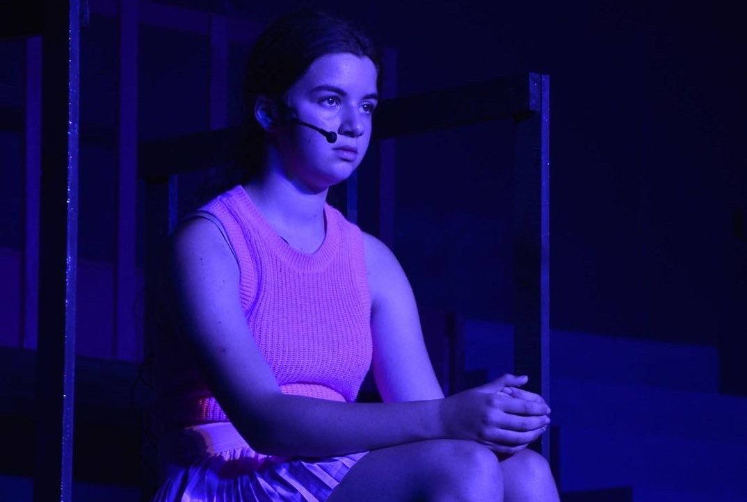 A person wearing a microphone headset and sitting on stage under blue lighting.