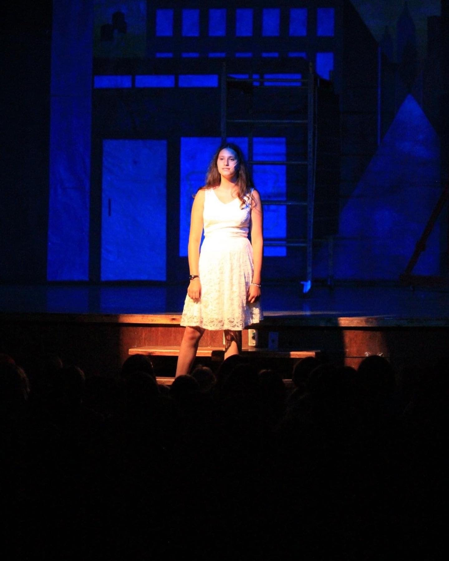 Young woman in a white dress standing on stage with blue lighting, audience in foreground.