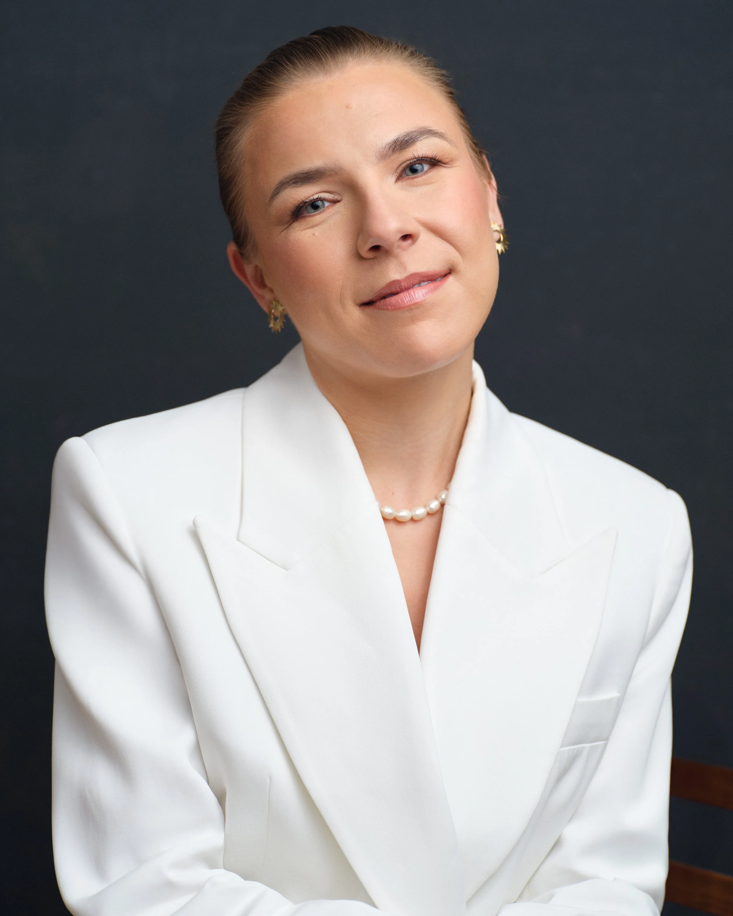 A woman with blond hair tied back, wearing a white suit jacket, pearl necklace, and gold earrings, smiling slightly against a dark gray background.