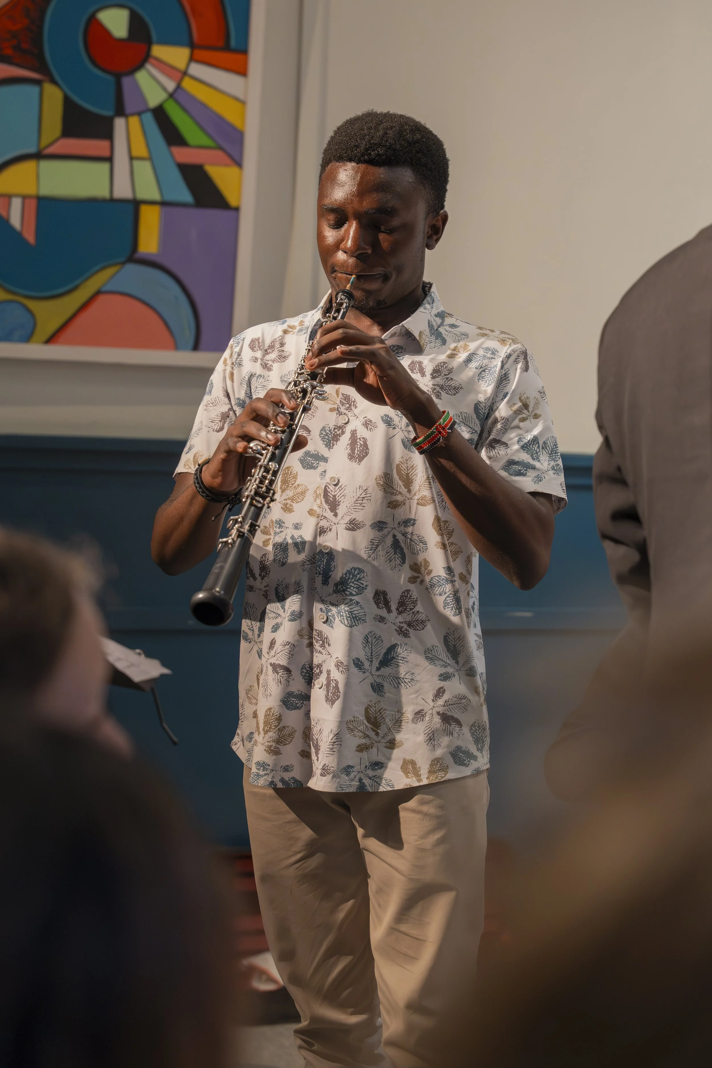 A young man in a short-sleeved shirt with a leaf pattern and beige pants playing a clarinet in an indoor setting with abstract colorful artwork on the wall behind him.