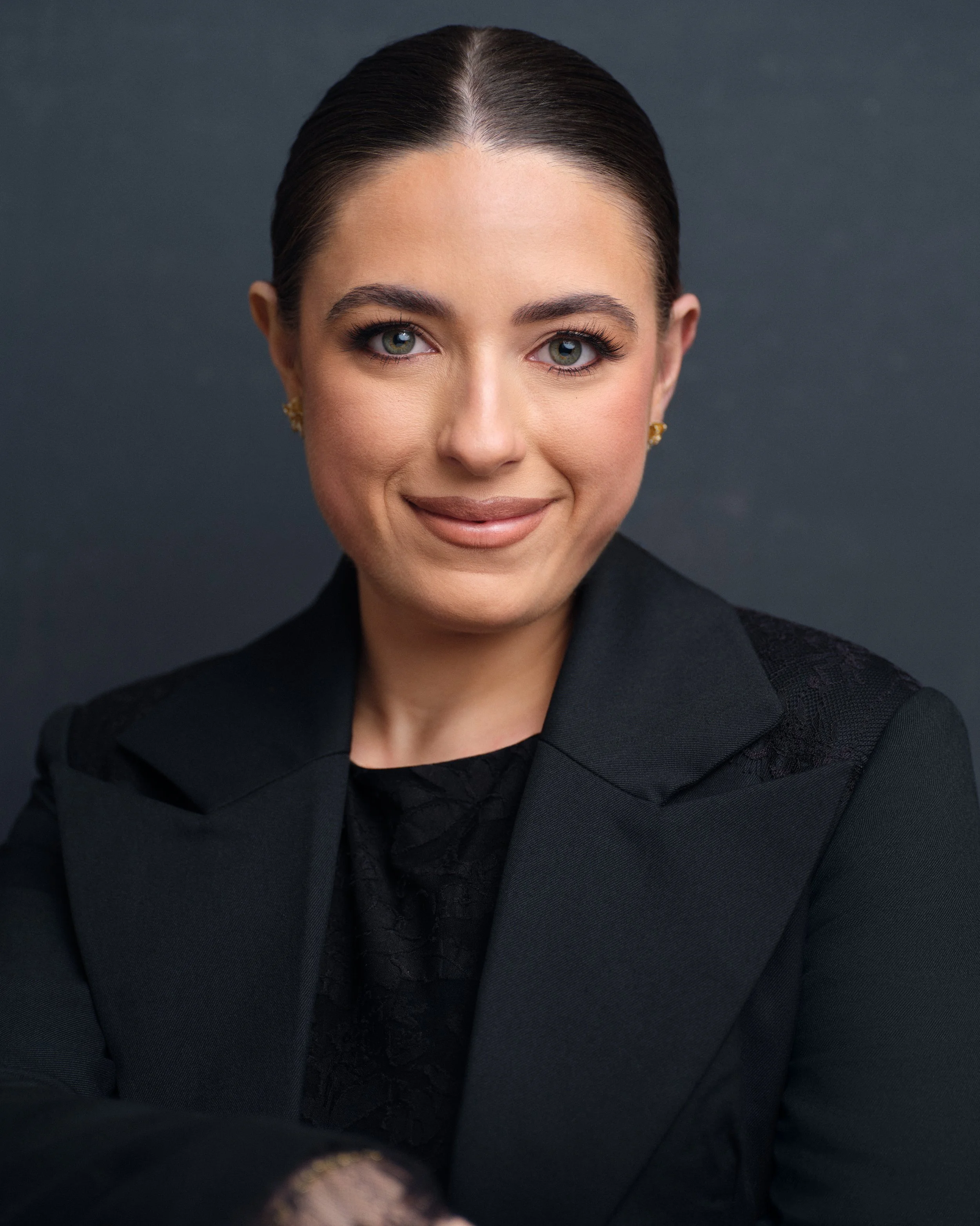 A young woman with dark hair and green eyes smiling while holding a violin in front of a gray background.