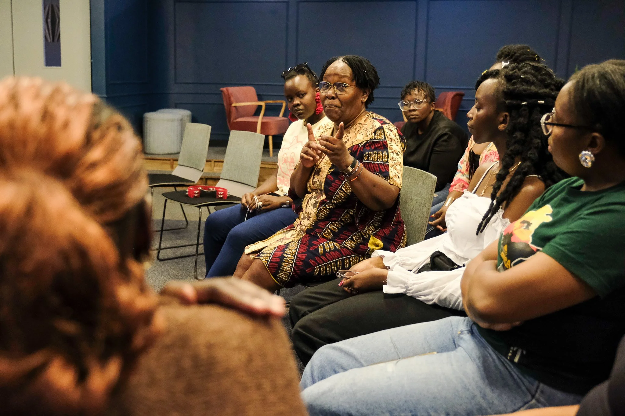 A group of women and girls sitting in chairs in a room with dark blue walls, engaged in a discussion or meeting. One woman in the center is speaking and gesturing with her hands, wearing glasses and a patterned dress. The others are listening attentively.