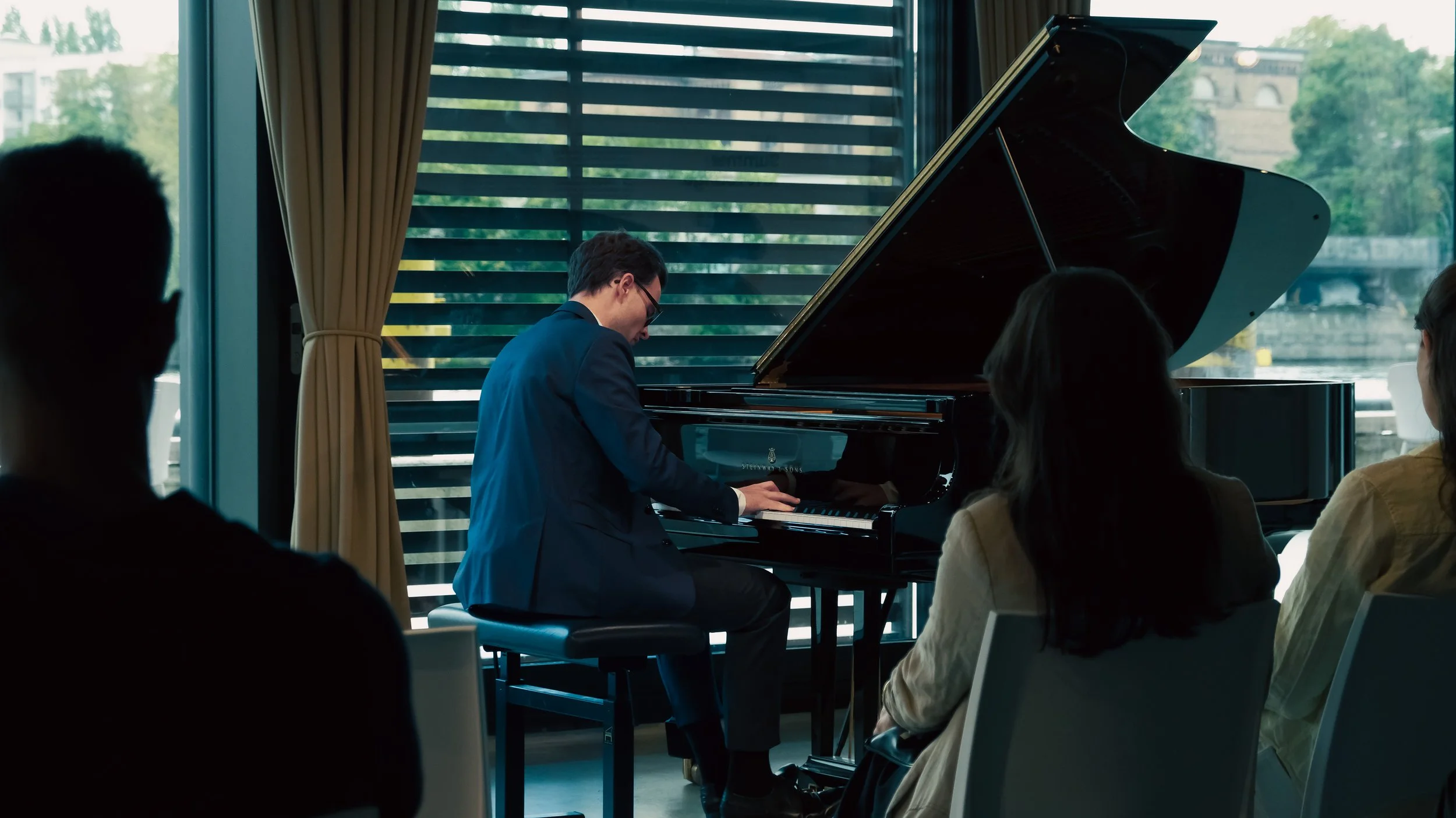 A person in a blue suit playing a grand piano during a performance or rehearsal, with an audience of three people watching silently inside a room with large windows showing trees and buildings outside.