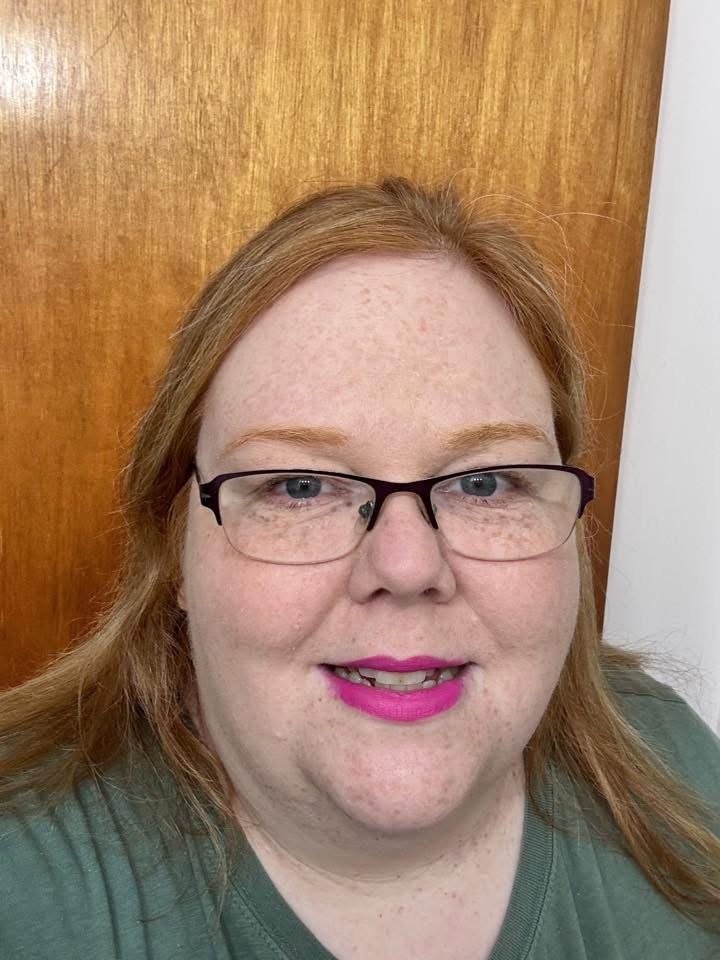 A woman with red hair, glasses, pink lipstick, and freckles smiling at the camera, standing in front of a wooden door.