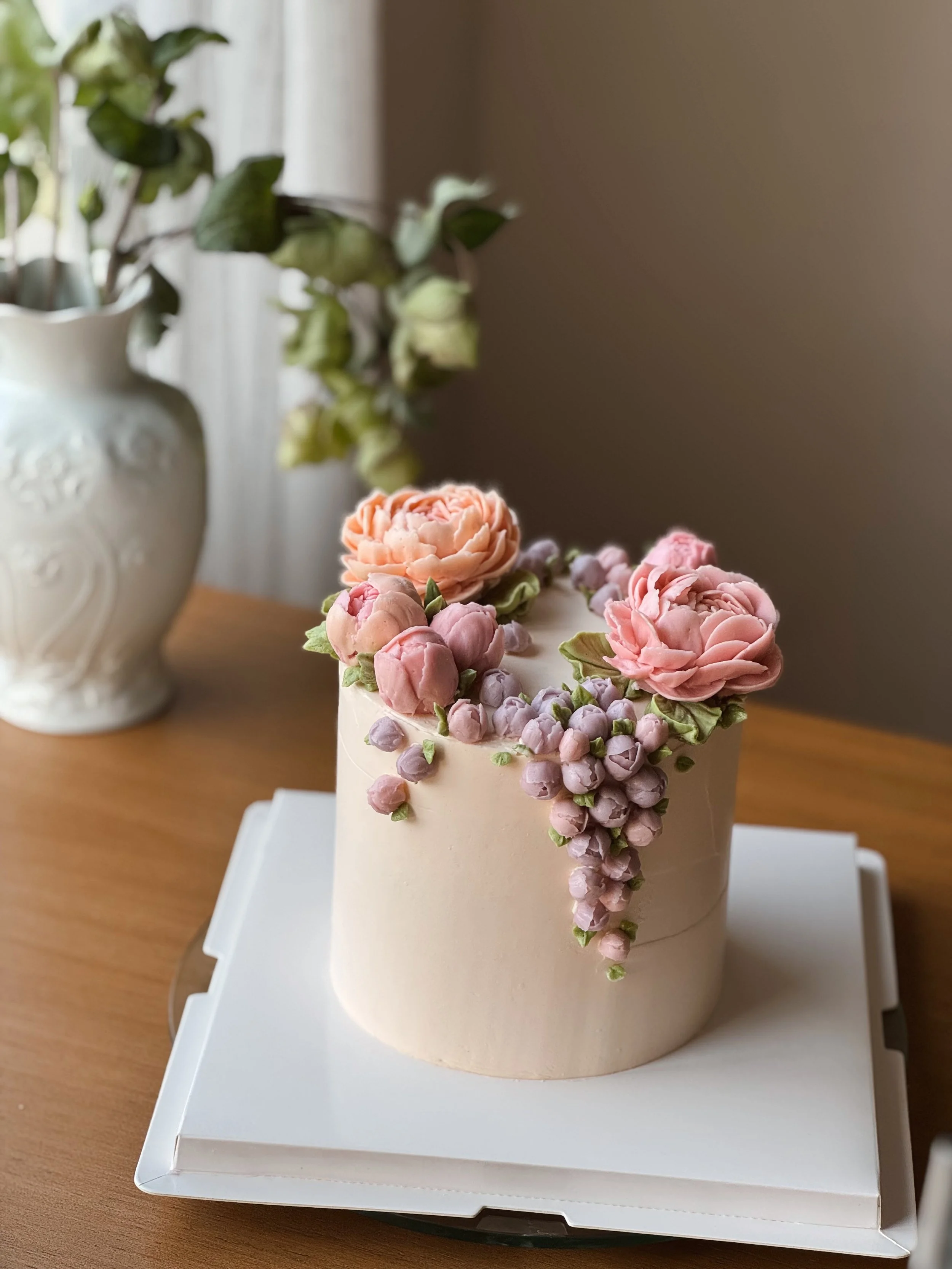 Pastel de cumpleaños decorado con flores de azúcar en tonos rosas y lilas, en una mesa de madera.