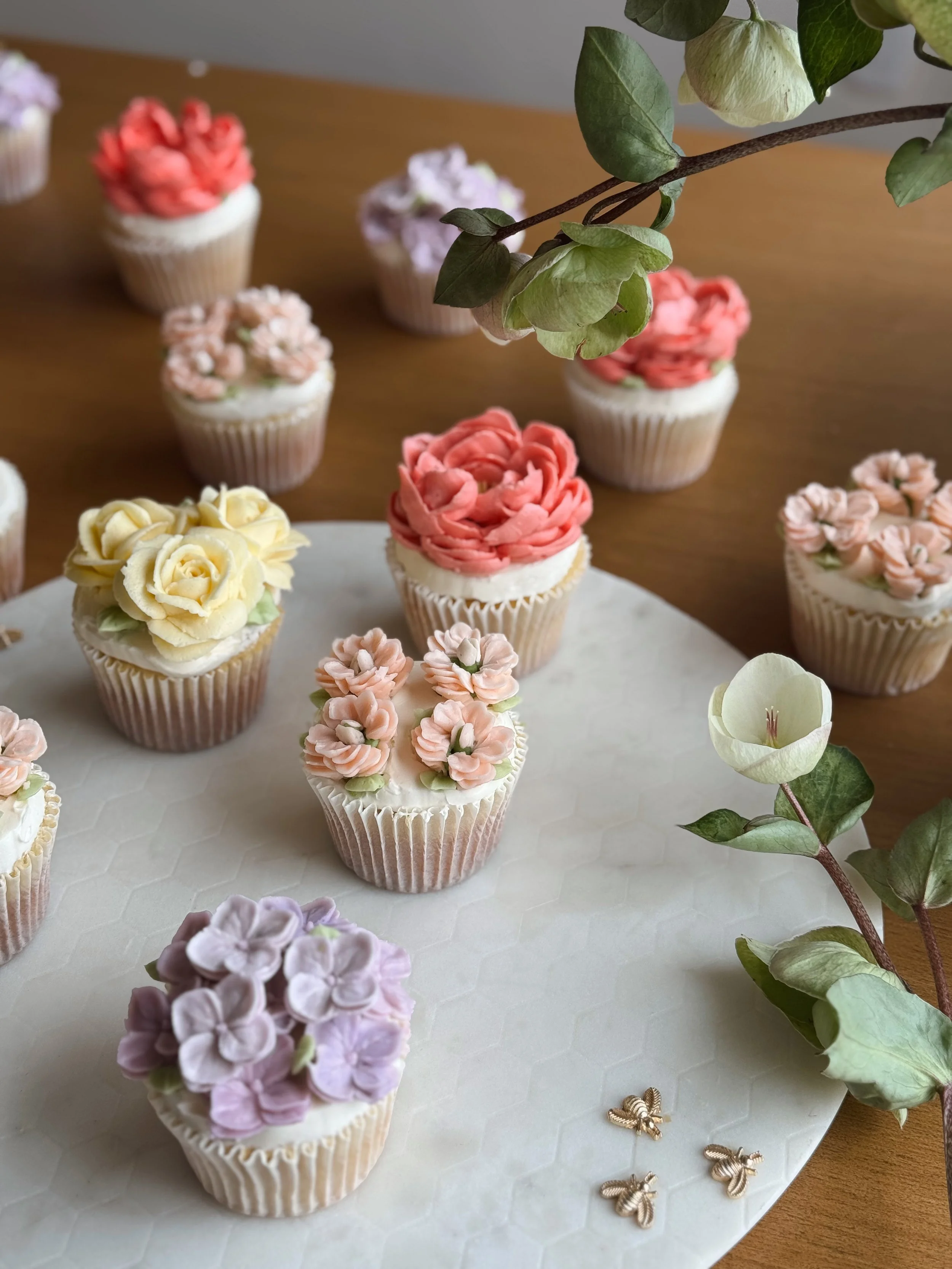 Un surtido de cupcakes decorados con flores de crema en colores amarillo, rosado, lavanda y beige, colocados sobre una base blanca, con hojas y flores decorativas, y pequeños accesorios en forma de abejas en la parte inferior derecha.