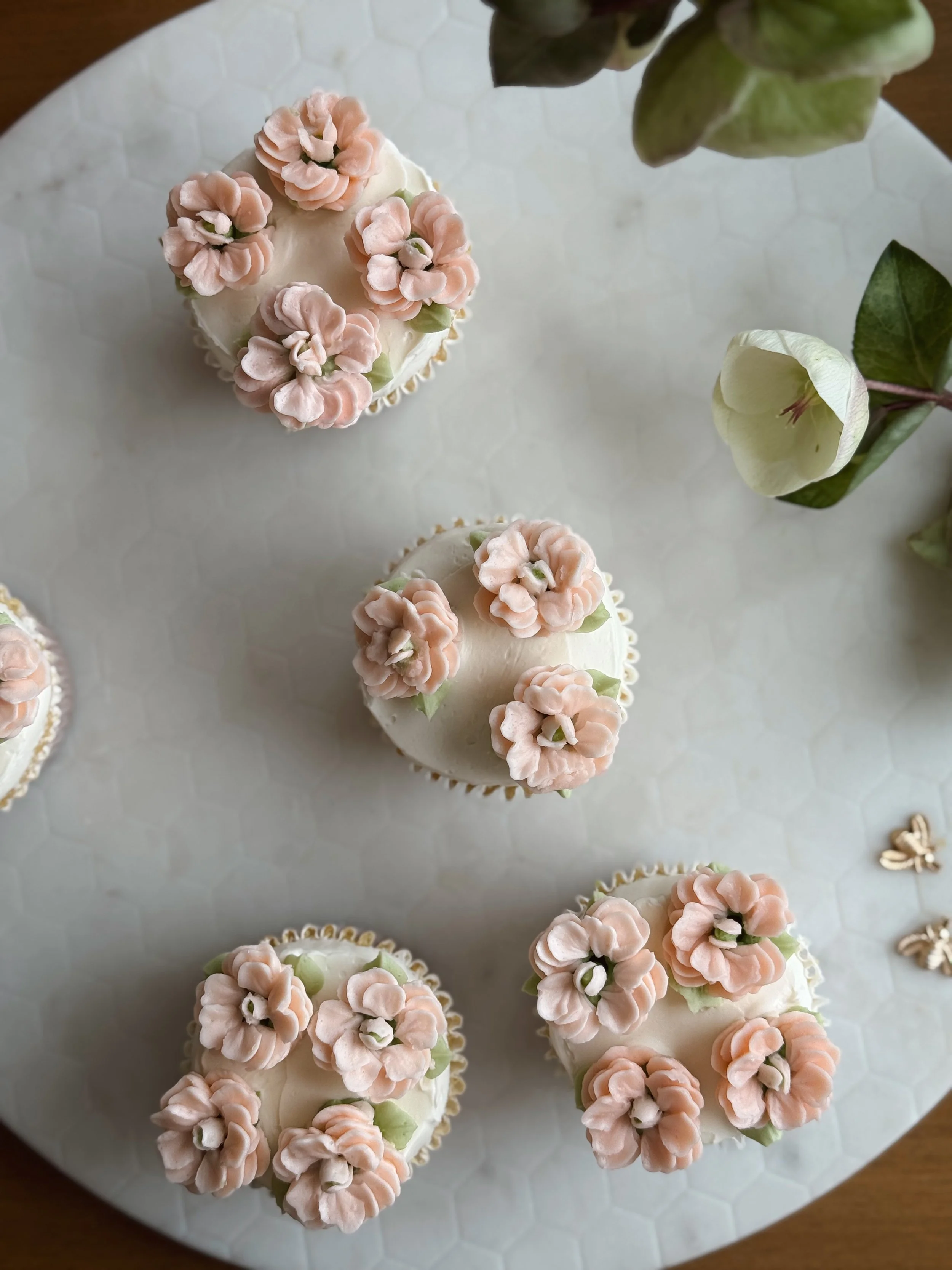 Cuatro cupcakes decorados con flores de color rosa en la parte superior en un plato de borde hexagonal, acompañado de hojas verdes y una flor blanca en el lado derecho.