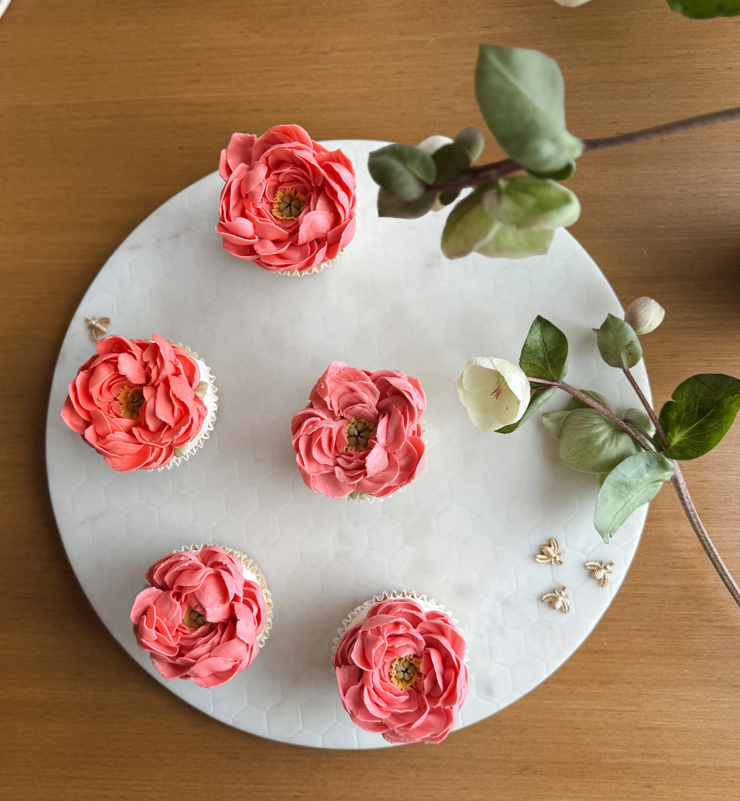 Seis cupcakes decorados con flores rosadas en un plato blanco en una superficie de madera y hojas verdes decorativas adjuntas.