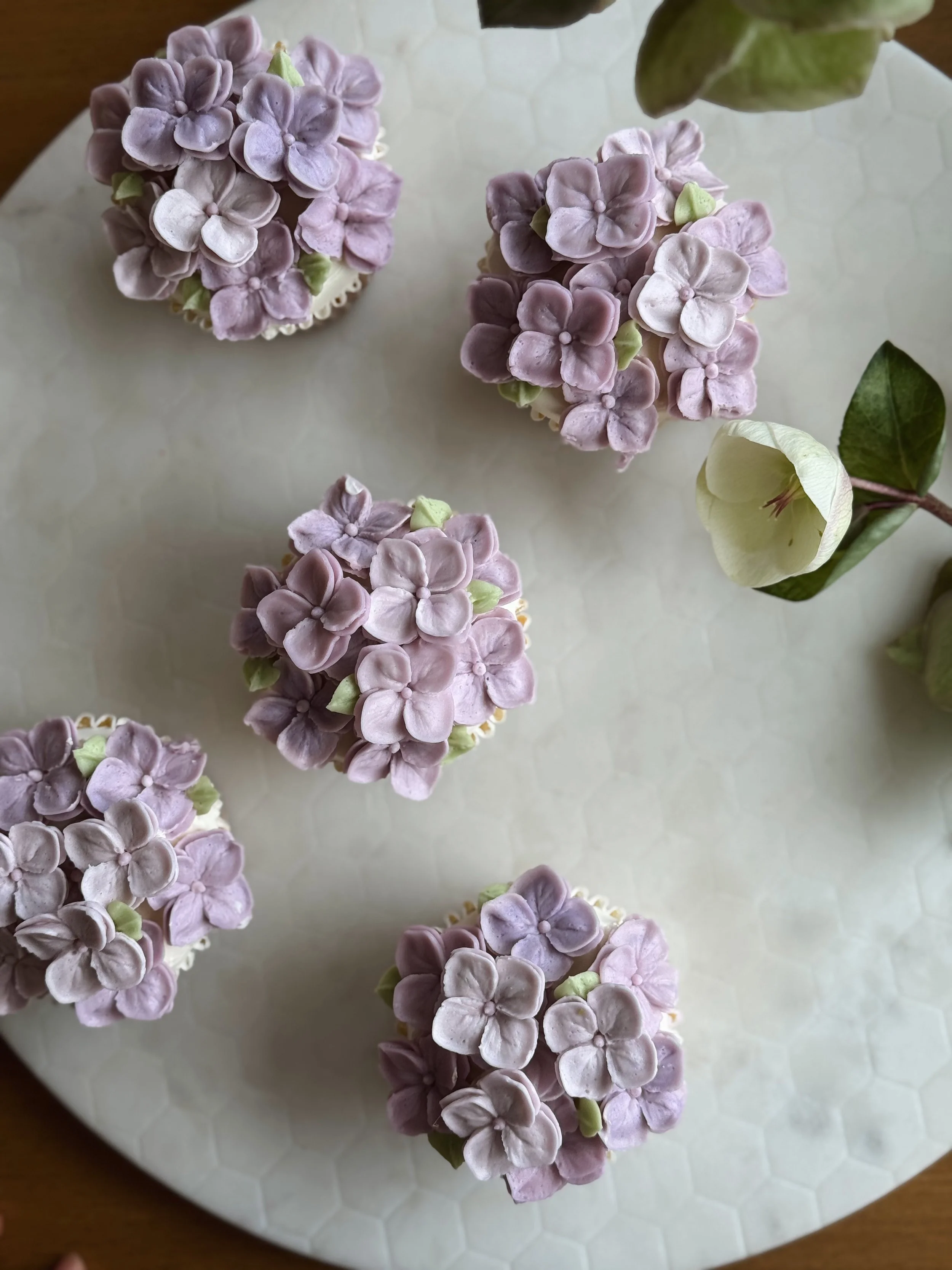 Cinco cupcakes decorados con flores de color rosa y morado, con detalles en verde, colocados en una bandeja blanca con superficie de patrón hexagonal, junto a una flor de papel o tela en tonos blanco y beige.