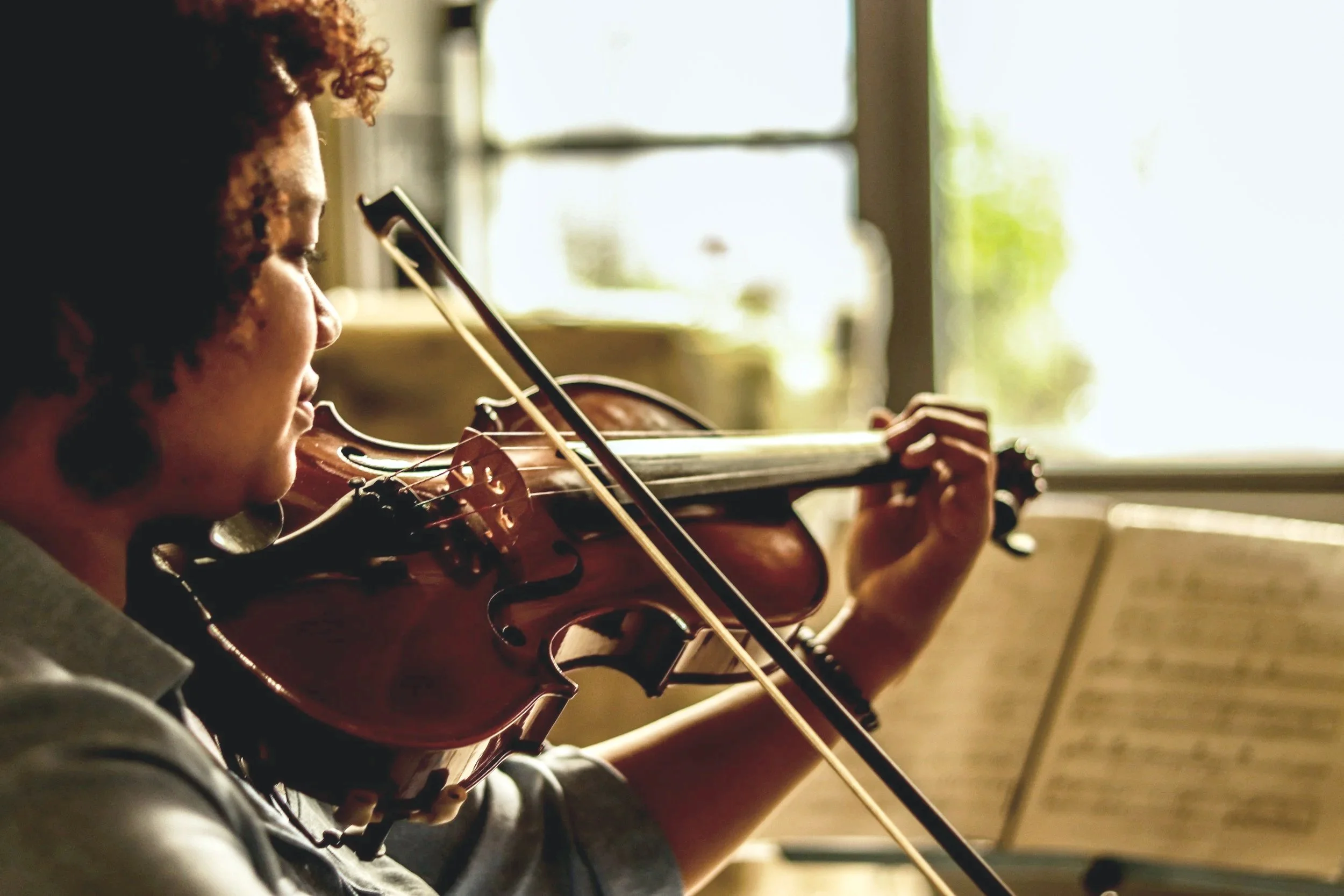 A woman playing a violin indoors near a window during daytime, with sheet music on a stand.